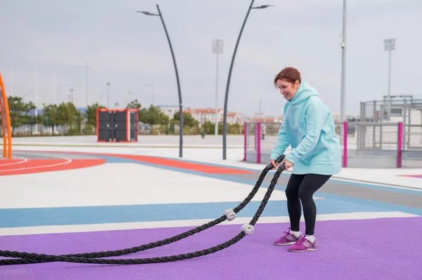 Caucasian woman in a mint sweatshirt is training with battle ropes at the sports ground