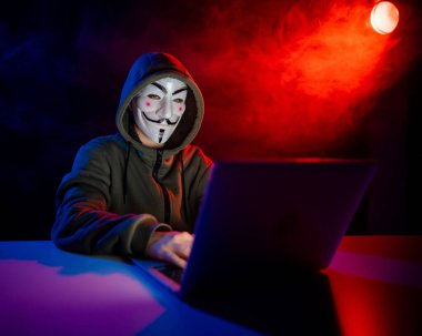 5 Haziran 2022 Novosibirsk, Rusya: Kapüşonlu Anonymous kırmızı-mavi dumanla karanlıkta dizüstü bilgisayarda yazıyor