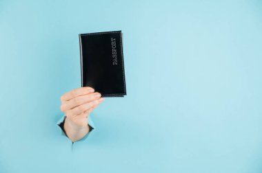 Bir kadının eli mavi bir kağıdın arkasındaki delikten dışarı çıkar ve siyah bir kapakta bir pasaport tutar.