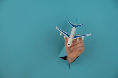 Mavi arka planda bir delikten dışarı sarkan bir kadın el bir uçak modeli tutar.