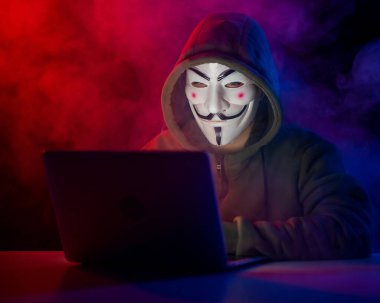 5 Haziran 2022 Novosibirsk, Rusya: Kapüşonlu Anonymous kırmızı-mavi dumanla karanlıkta dizüstü bilgisayarda yazıyor