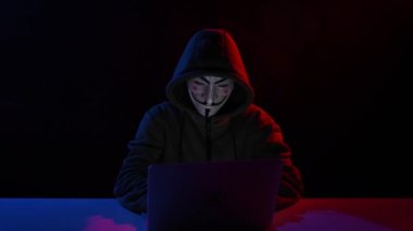 5 Haziran 2022 Novosibirsk, Rusya: Kapüşonlu Anonymous kırmızı ışıkta karanlıkta dizüstü bilgisayarda yazıyor