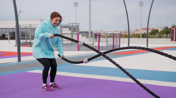 Caucasian woman in a mint sweatshirt is training with battle ropes at the sports ground