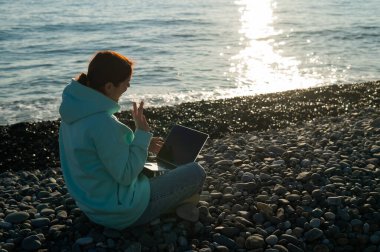 Kumsalda otururken bilgisayardaki video bağlantısıyla konuşan beyaz bir kadın.