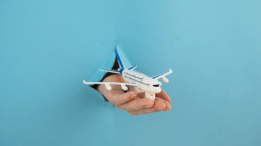 Mavi arka planda bir delikten dışarı sarkan bir kadın el bir uçak modeli tutar.