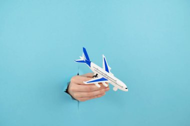 Mavi arka planda bir delikten dışarı sarkan bir kadın el bir uçak modeli tutar.