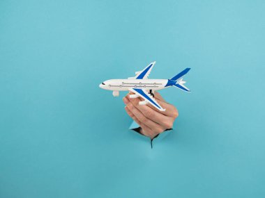 Mavi arka planda bir delikten dışarı sarkan bir kadın el bir uçak modeli tutar.