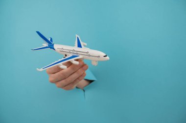 Mavi arka planda bir delikten dışarı sarkan bir kadın el bir uçak modeli tutar.