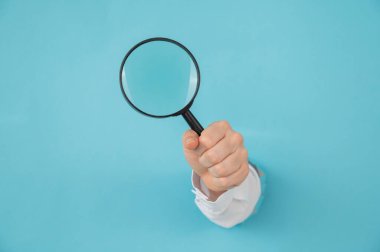 Bir kadının eli kağıtta bir delikten dışarı sarkan mavi arka planda büyüteç tutar.