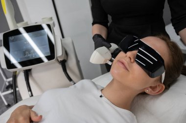 Gözlüklü genç beyaz kadın, fotojenizasyon prosedürü uyguluyor. Güzellik kliniğindeki bir kızın portresi.