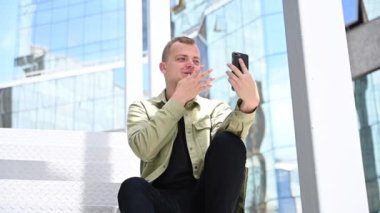 Genç adam merdivenlerde oturup dışarıdaki akıllı telefondan işaret diliyle konuşuyor.