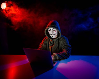 5 Haziran 2022 Novosibirsk, Rusya: Kapüşonlu Anonymous kırmızı-mavi dumanla karanlıkta dizüstü bilgisayarda yazıyor