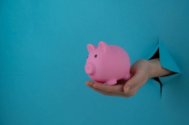 Mavi arka planda bir delikten dışarı sarkan bir kadın eli pembe bir kumbara tutuyor.