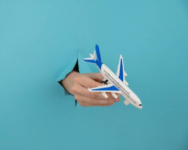 Mavi arka planda bir delikten dışarı sarkan bir kadın el bir uçak modeli tutar.