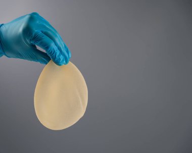Eldivenli bir kadın eli gri arka planda gözyaşı şeklinde bir göğüs implantı tutuyor.
