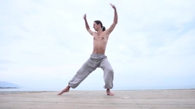 Beyaz bir adam, çıplak gövdesiyle deniz kıyısında wushu yapıyor.