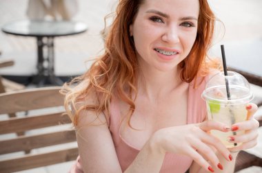 Diş teli takan güzel, kızıl saçlı bir kadın yazın dışarıda soğuk limonata içiyor. Çilli, gülümseyen bir kızın portresi.