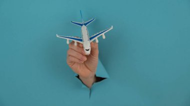 Mavi arka planda bir delikten dışarı sarkan bir kadın el bir uçak modeli tutar.