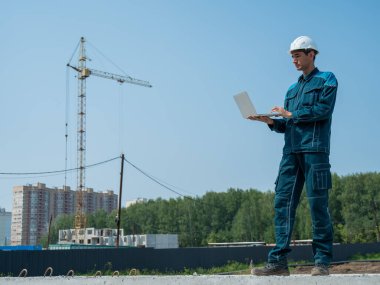 Kasklı bir erkek müteahhit elinde dizüstü bilgisayarla inşaat alanının arka planında duruyor.