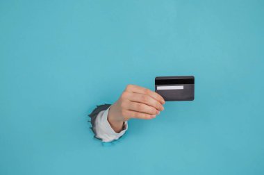Bir kadının eli kağıtta bir delikten dışarı sarkan mavi arka planda bir kredi kartı tutar.