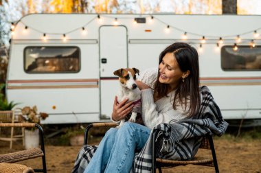 Beyaz kadın hasır bir sandalyede vagonun yanında oturuyor ve dışarıda Jack Russell Terrier 'ın köpeğine sarılıyor. Sonbaharda bir karavanla seyahat et.