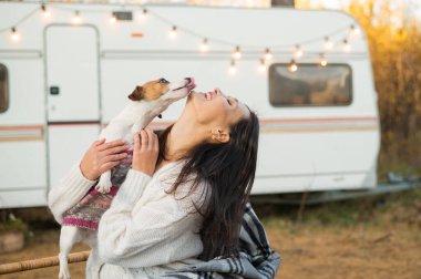 Beyaz kadın hasır bir sandalyede vagonun yanında oturuyor ve dışarıda Jack Russell Terrier 'ın köpeğine sarılıyor. Sonbaharda bir karavanla seyahat et.