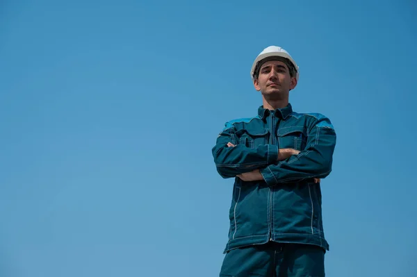 A caucasian man in work clothes and a construction helmet stands against the blue sky with his arms crossed on his chest
