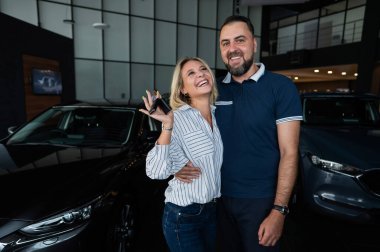 Mutlu beyaz çift kucaklaşıyor ve kadın araba galerisinde yeni araba anahtarlarını tutuyor.