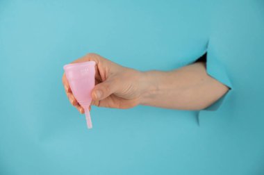 Mavi arka planda bir delikten dışarı sarkan bir kadın eli pembe bir adet bardağı tutuyor.