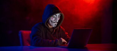 5 Haziran 2022 Novosibirsk, Rusya: Kapüşonlu Anonymous kırmızı-mavi dumanla karanlıkta dizüstü bilgisayarda yazıyor