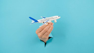 Mavi arka planda bir delikten dışarı sarkan bir kadın el bir uçak modeli tutar.