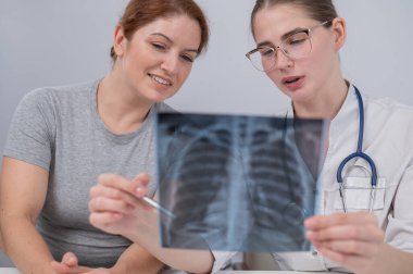 Doktor beyaz bir kadına danışıyor ve akciğerlerin röntgenleri hakkında yorum yapıyor.