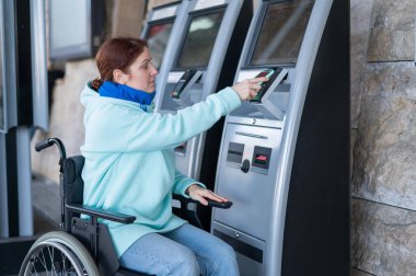 Tekerlekli sandalyedeki beyaz bir kadın, bir self-servis kasasında cep telefonu kullanarak tren bileti alıyor.