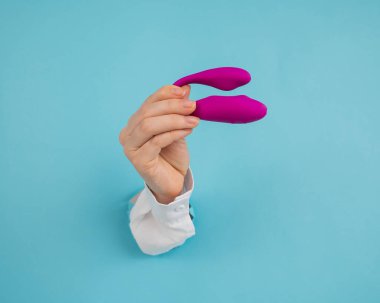 Bir kadının eli pembe bir seks oyuncağını kağıt mavisi bir arka planda bir delikten tutar.