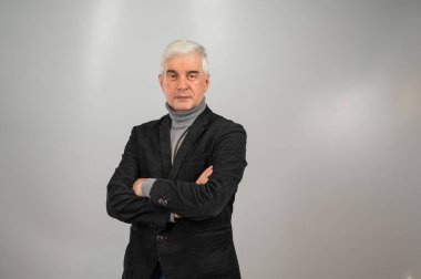 Yaşlı, gri saçlı, takım elbiseli bir adam kollarını göğsünde kavuşturmuş duruyor.