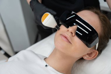 Gözlüklü genç beyaz kadın, fotojenizasyon prosedürü uyguluyor. Güzellik kliniğindeki bir kızın portresi.