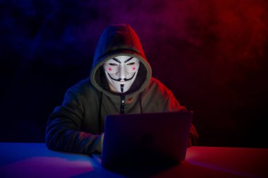 5 Haziran 2022 Novosibirsk, Rusya: Kapüşonlu Anonymous kırmızı-mavi dumanla karanlıkta dizüstü bilgisayarda yazıyor