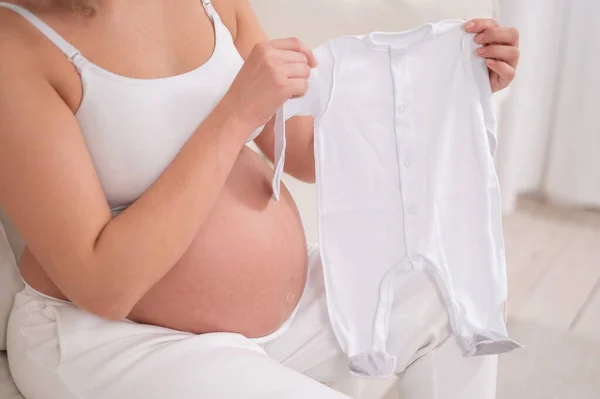 妊娠中の女性は白いソファに座って 予想される赤ちゃんのために服を保持します — ストック写真