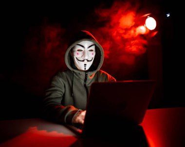 Kapüşonlu anonim, kırmızı dumanla karanlıkta dizüstü bilgisayarda yazıyor.