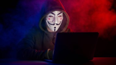 5 Haziran 2022 Novosibirsk, Rusya: Kapüşonlu Anonymous kırmızı-mavi dumanla karanlıkta dizüstü bilgisayarda yazıyor