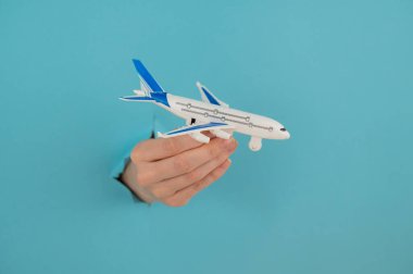 Mavi arka planda bir delikten dışarı sarkan bir kadın el bir uçak modeli tutar.