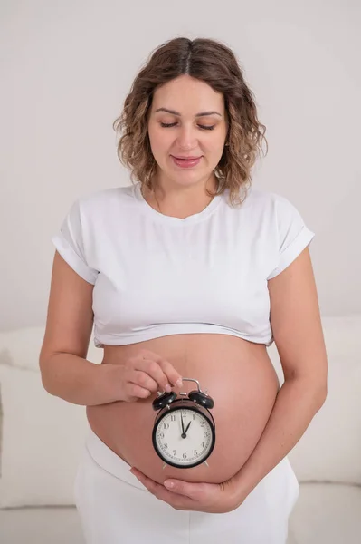 裸の腹を持つ妊婦は目覚まし時計を持っている 出生の予定日 — ストック写真