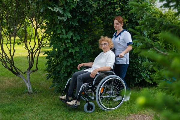 白人女医生和一位坐在轮椅上的老年病人在公园里散步 护士陪着一位老妇人在户外散步 — 图库照片