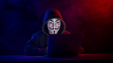5 Haziran 2022 Novosibirsk, Rusya: Kapüşonlu Anonymous kırmızı-mavi dumanla karanlıkta dizüstü bilgisayarda yazıyor