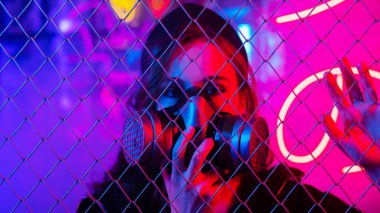 Neon bir stüdyoda çitin arkasında gaz maskeli beyaz bir kadın.