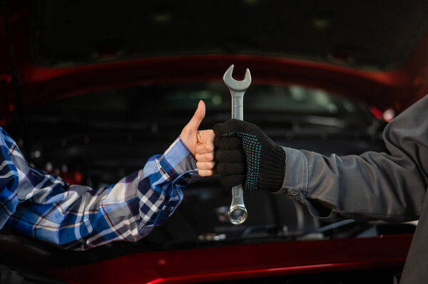 Close-up of womans hand thumb up and mechanics hand with tool. Automotive master in gloves shows a positive gesture after finishing work.