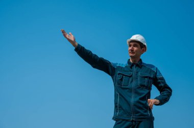 İş kıyafetleri içinde beyaz bir adam ve bir miğfer kollarını yana açmış mavi bir gökyüzünün arka planında duruyor.