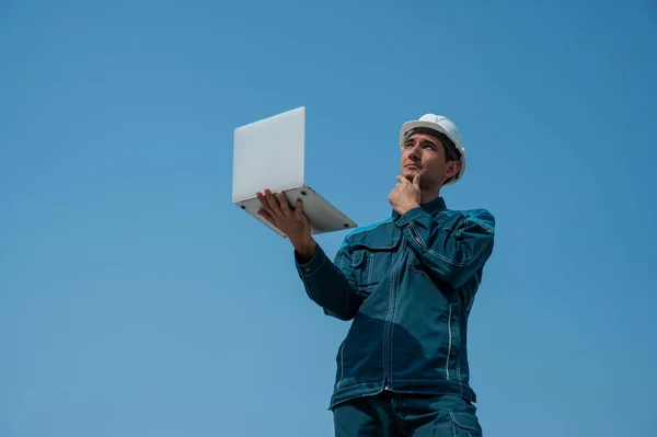 ヘルメットを被った男が青い空に向かって立っているノートパソコンを手に — ストック写真