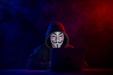 5 Haziran 2022 Novosibirsk, Rusya: Kapüşonlu Anonymous kırmızı-mavi dumanla karanlıkta dizüstü bilgisayarda yazıyor