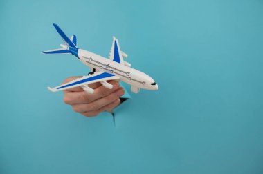Mavi arka planda bir delikten dışarı sarkan bir kadın el bir uçak modeli tutar.